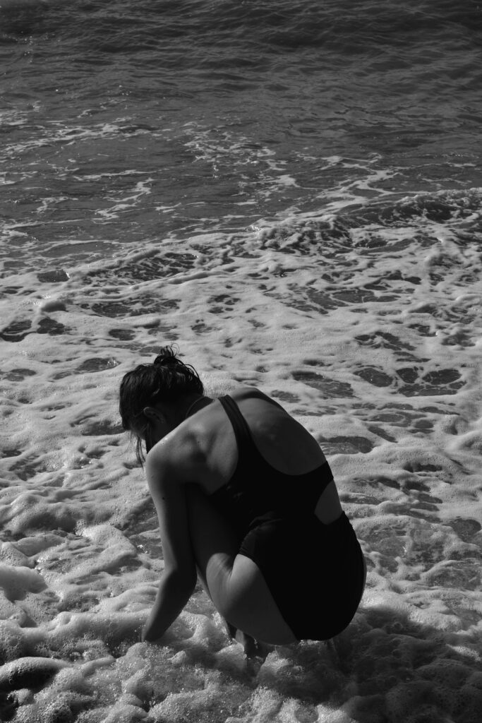 A woman in swimwear crouches by the ocean shore, surrounded by foamy waves.