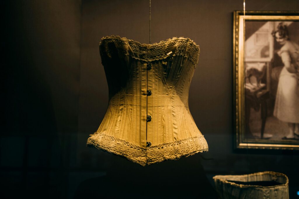 pexels-photo-5326766-5326766 Bright historical corset with buttons hanging on cord in showcase near picture in museum
